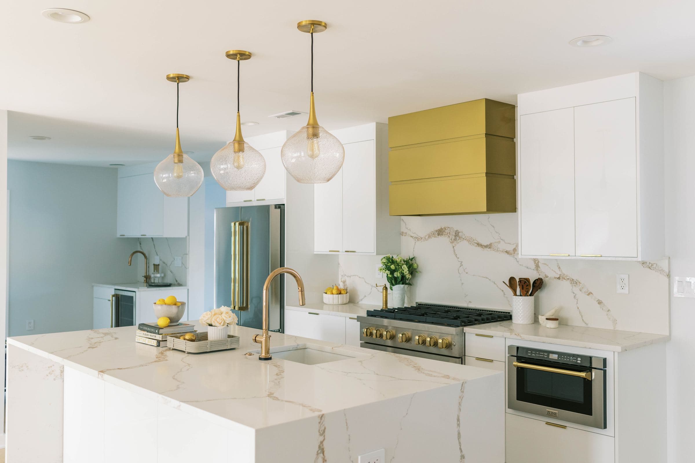 Angled view of a white kitchen with a brass hood and veined waterfall island